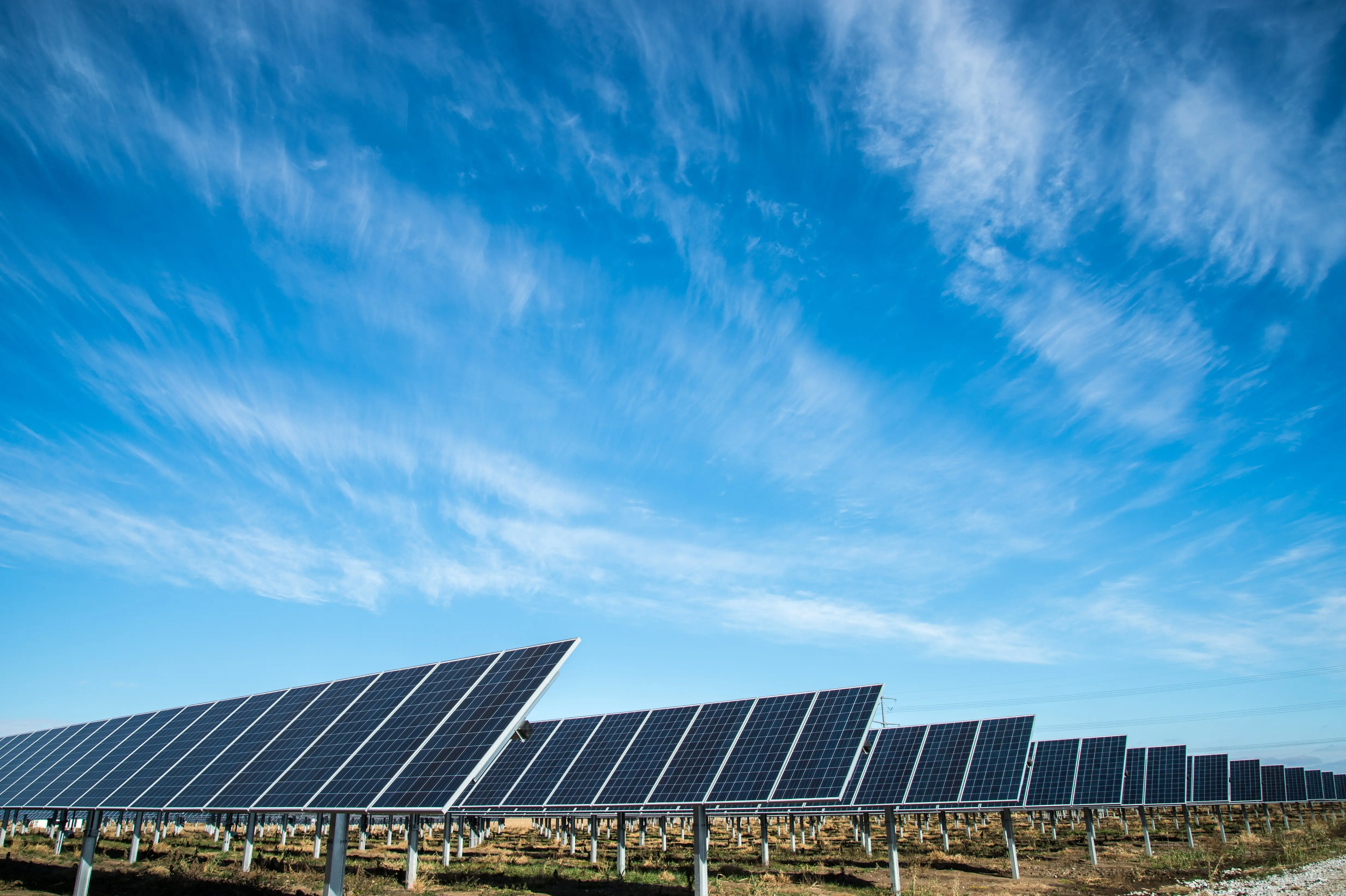 Ein Solarpark mit mehreren Reihen von Photovoltaik-Solarmodulen, die auf Metallstrukturen montiert sind, unter einem weiten blauen Himmel mit dünnen, zarten Wolken. Die Anlage erzeugt erneuerbare Energie und befindet sich in einer flachen, offenen Landschaft bei Tageslicht.