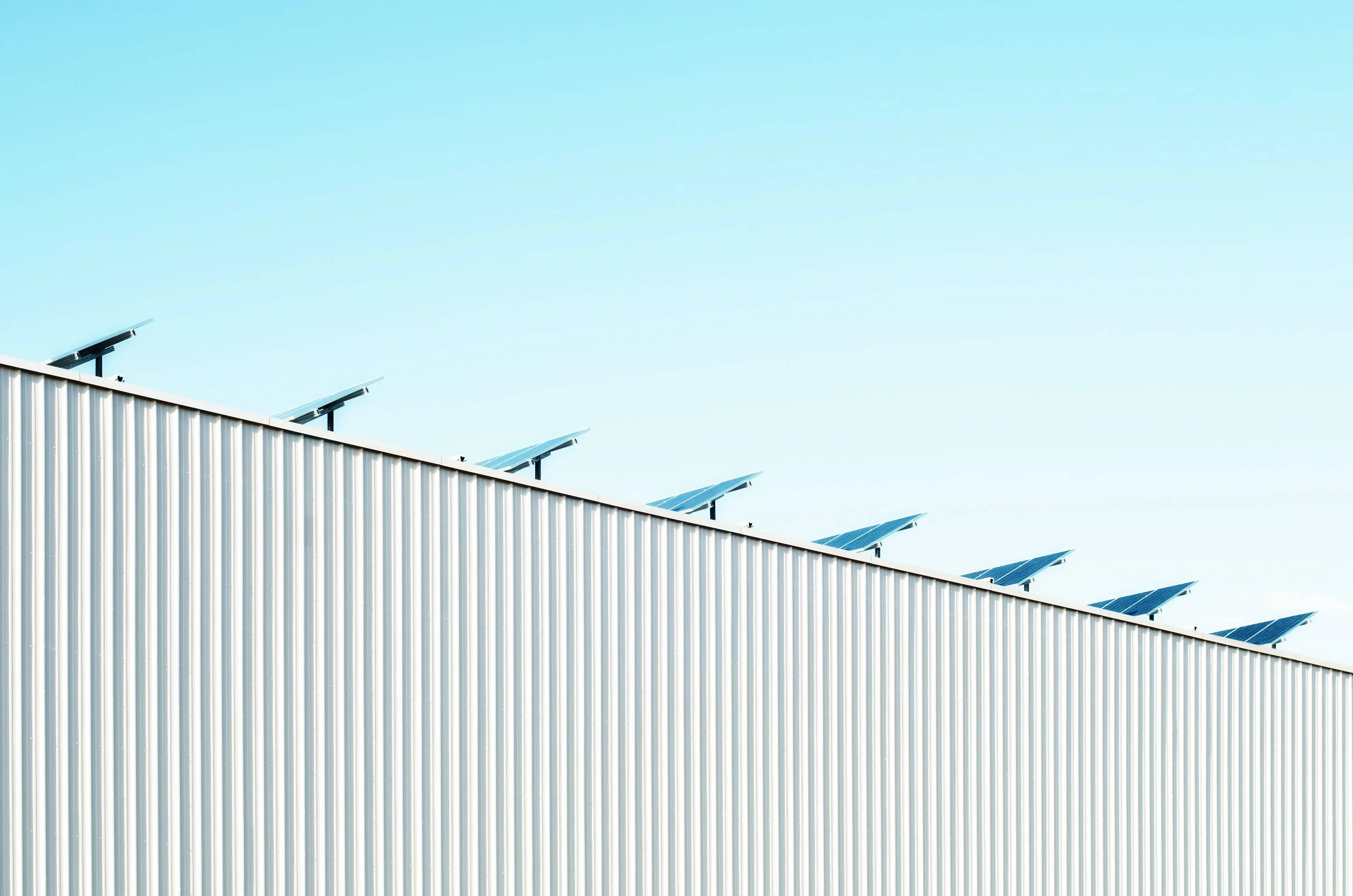 Eine Reihe von Solarpanelen, die auf dem Dach eines Gebäudes mit vertikaler Metallverkleidung installiert sind. Die Solarpanele sind in gleichmäßigen Abständen aufgereiht und zeigen alle in die gleiche Richtung. Der Himmel im Hintergrund ist klar und blau, was auf gutes Wetter hinweist. Die Szene symbolisiert erneuerbare Energie und moderne Nachhaltigkeit.
