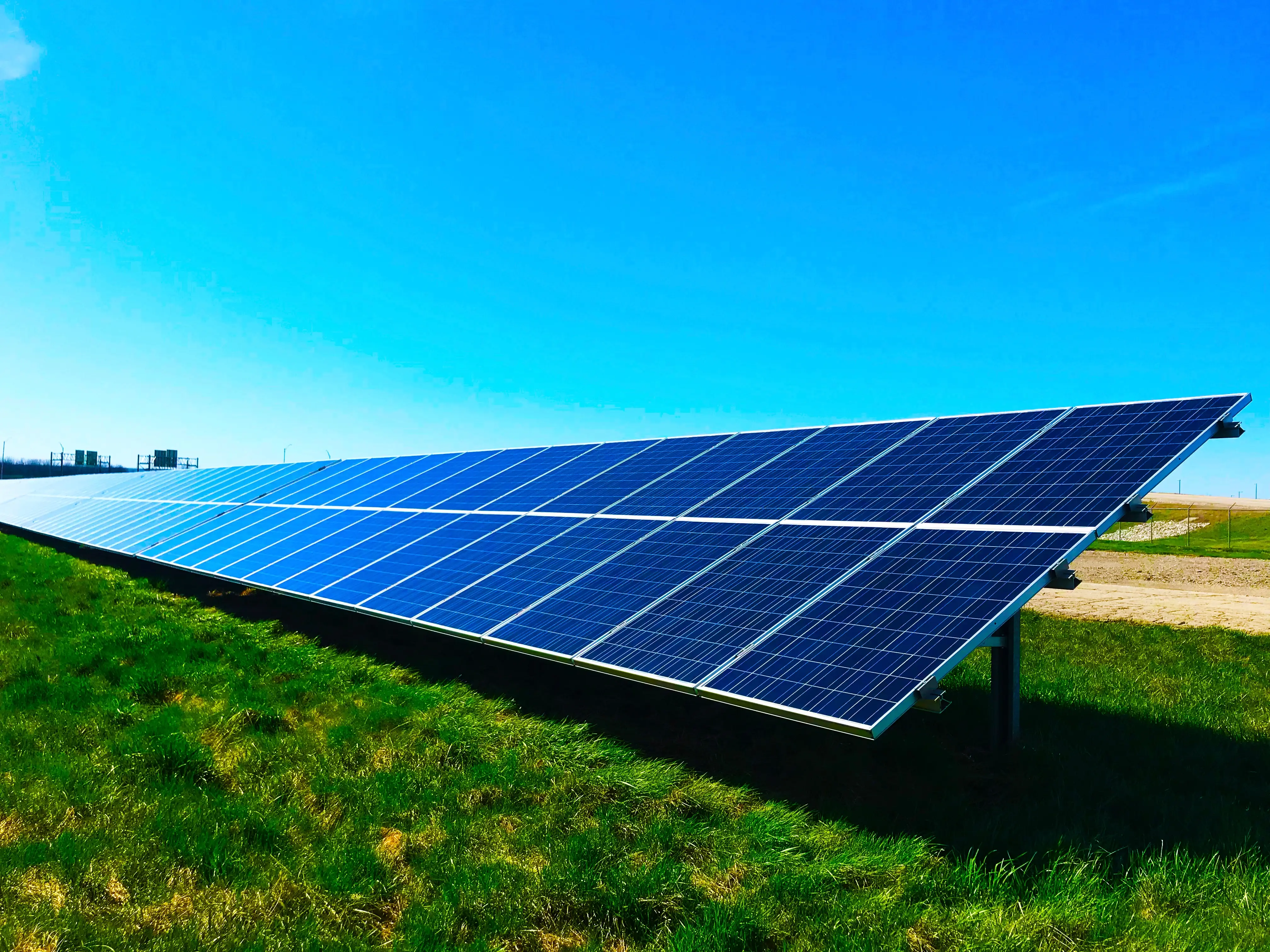 Ein großes Feld von Photovoltaik-Solarmodulen unter einem klaren blauen Himmel. Die Module sind in einer Neigung montiert, um die Sonneneinstrahlung optimal zu nutzen. Unter den Paneelen ist grünes Gras sichtbar.
